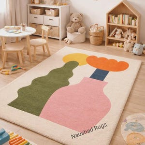 Può includere: Un tappeto rettangolare con sfondo color crema caratterizzato da un grande vaso rosa, uno stelo blu scuro, un fiore arancione, un sole giallo e una forma verde ondulata. Il testo Naushad Rugs è stampato in nero sul lato inferiore destro.