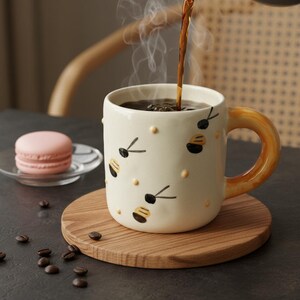 Può includere: Una tazza in ceramica color crema con manico giallo e decorazioni a forma di api. Il caffè viene versato nella tazza, che poggia su un sottobicchiere di legno. Un macaron rosa è su un piattino di vetro. Chicchi di caffè sparsi.