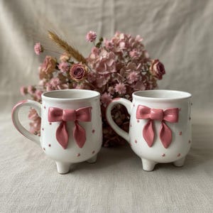 Puede incluir: Dos tazas de cer&aacute;mica blancas con lazos rosas y peque&ntilde;os lunares rosas. Cada taza tiene un asa y se apoya en tres patas peque&ntilde;as. Flores rosas secas en el fondo.