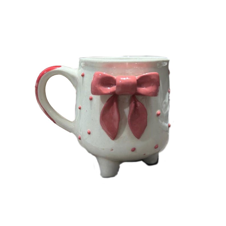 Puede incluir: Taza de cer&aacute;mica blanca con un lazo rosa y lunares. La taza tiene un asa rosa y est&aacute; sostenida por tres peque&ntilde;os pies. El lazo es un elemento decorativo prominente en la parte delantera de la taza.