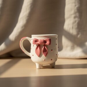 Puede incluir: Una taza de cer&aacute;mica blanca con un lazo rosa y peque&ntilde;os lunares rosas. La taza tiene un asa curvada y se apoya en tres peque&ntilde;os pies. El lazo es un elemento decorativo prominente en la parte delantera de la taza.