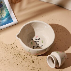May include: Cream ceramic matcha bowl with a spout, inscribed with "I Love you so MATCHA" and floral details. A matching whisk holder is next to the bowl. Green matcha powder is scattered around.