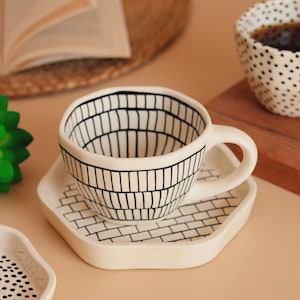 Puede incluir: Un juego de taza y platillo de café de cerámica blanca. La taza presenta un patrón de ladrillos negros en el exterior, con un patrón a juego en el platillo hexagonal. En el fondo se ve una segunda taza con lunares negros.