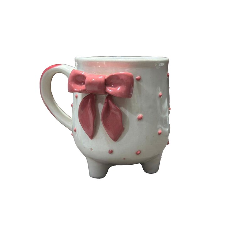 Puede incluir: Taza de cer&aacute;mica blanca con un lazo rosa y lunares rosas. La taza tiene un asa curva y se apoya en tres peque&ntilde;os pies. El lazo es un elemento decorativo prominente, que a&ntilde;ade un toque de encanto al dise&ntilde;o. La taza es un art&iacute;culo decorativo.