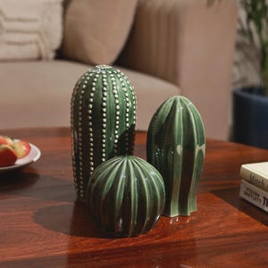 May include: Three green ceramic cactus ornaments of different shapes and sizes. The tallest is cylindrical with white dot detailing. The others are a sphere and a ridged, upright form. They are displayed on a dark wooden table.