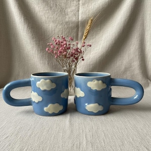 Puede incluir: Dos tazas de cerámica azules con diseños de nubes blancas. Cada taza tiene un asa grande y curvada y está decorada con nubes blancas y esponjosas. Las tazas están colocadas sobre una superficie de color neutro.
