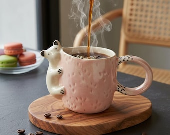 May include: A pink ceramic mug with a white polar bear design, filled with coffee. The mug has a textured surface and a handle with black spots. The coffee is being poured from a carafe, and steam rises from the mug. The mug sits on a wooden coaster.