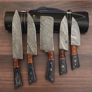 Könnte beinhalten: Ein Set aus fünf Damaststahlmessern mit Holz- und schwarzen Griffen, präsentiert vor einer schwarzen Leder-Messertasche. Die Klingen haben ein einzigartiges, gemustertes Design. Die Griffe sind eine Kombination aus Holz und schwarzem Material.