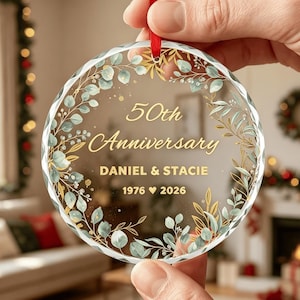 Könnte beinhalten: Ein rundes, transparentes Acryl-Ornament mit einem goldfarbenen Kranz aus Eukalyptusblättern. Der Text lautet 50th Anniversary, DANIEL & STACIE und 1976 Herz 2026. Das Ornament ist an einem dünnen roten Band zum Aufhängen befestigt.