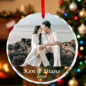 Könnte beinhalten: Ein runder Weihnachtsbaumschmuck aus Glas mit einem Foto von zwei Personen, die auf Felsen am Meer sitzen. Der Anhänger hat ein rotes Band und ist unten mit dem weißen Text Ken & Diane sowie der Jahreszahl 2026 bedruckt.
