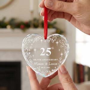 K&ouml;nnte beinhalten: Ein herzf&ouml;rmiges Ornament aus klarem Glas mit facettierten Kanten, verziert mit einem wei&szlig;en, ge&auml;tzten Blumenkranz-Design, dem Text 25th SILVER ANNIVERSARY, den Namen Mason & Louise und dem Datum 09.21.2001, aufgeh&auml;ngt an einem roten Band.