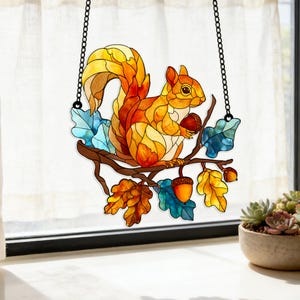 Peut inclure: Suncatcher en vitrail représentant un écureuil orange et jaune sur une branche marron, tenant un gland. Les feuilles sont bleues et jaunes. Suspendu par une chaîne noire.