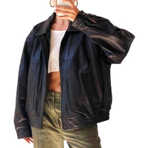 Könnte beinhalten: Schwarze Lederjacke mit Reißverschluss, Kragen und geripptem Saum. Die Jacke hat ein klassisches Design mit leicht übergroßer Passform. Die Ärmel sind lang und haben geknöpfte Manschetten. Die Jacke wird mit einem weißen Oberteil und olivgrünen Hosen getragen.