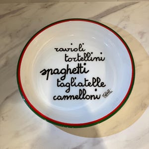 Pode incluir: Tigela de massa de cerâmica branca com borda vermelha e verde. A tigela apresenta as palavras "ravioli, tortellini, spaghetti, tagliatelle e cannelloni" em escrita preta. Uma assinatura é visível no lado direito da tigela.