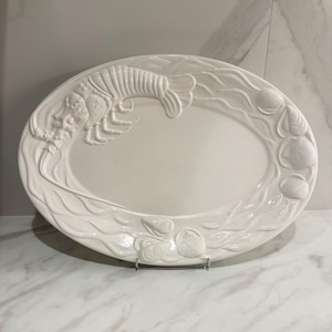 Może przedstawiać: Owalny, kremowy półmisek do serwowania z reliefowym wzorem. Wzór przedstawia homara i muszle, z falistymi wzorami na krawędzi. Półmisek jest podparty metalowym stojakiem.