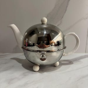 Può includere: Una teiera sferica con corpo in cromo lucido e beccuccio, manico, pomello del coperchio e tre piccoli piedini sferici in ceramica bianca. Il centro della teiera include un meccanismo di chiusura in metallo e la superficie riflette la stanza.