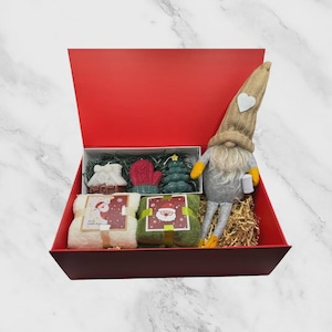 Könnte beinhalten: Eine rote Geschenkbox gefüllt mit weihnachtlichen Artikeln. Enthält einen Wichtel, zwei kleine Handtücher mit Weihnachtsmann-Motiven und dekorative Seifen in Form eines Hauses, Fäustlingen und eines Weihnachtsbaums. Die Box ist mit dekorativem Stroh ausgekleidet.