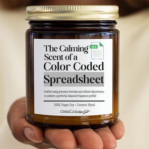 Könnte beinhalten: Ein braunes Glas-Kerzenglas mit goldenem Deckel. Auf dem Etikett steht "The Calming Scent of a Color Coded Spreadsheet". Die Kerze wird aus einer veganen Soja- und Kokosnussmischung hergestellt.