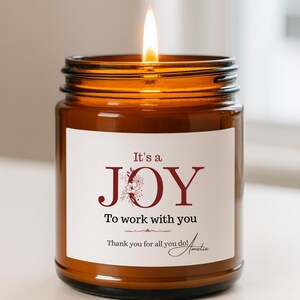 Può includere: Una candela accesa in un barattolo di vetro ambrato con un'etichetta bianca. L'etichetta dice "It's a JOY To work with you" in testo bordeaux, con un disegno floreale. La candela ha una fiamma luminosa.