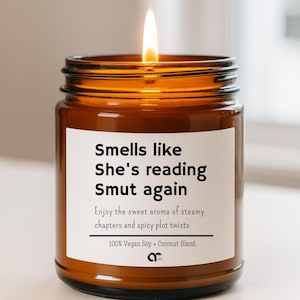 May include: A lit candle in a brown glass jar with a white label. The label reads "Smells like She's reading Smut again." The text below says "Enjoy the sweet aroma of steamy chapters and spicy plot twists." The candle is a 100% Vegan Soy + Coconut Blend.