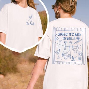 May include: Off-white t-shirt with blue graphic. Front features a cocktail glass and text "The Bride". Back has "CHARLOTTE'S BACH KEY WEST, FL" with bikini, seashell, and date graphics. A fun, casual tee.