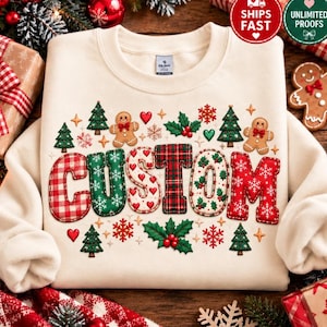 May include: A cream-colored crewneck sweatshirt featuring the word CUSTOM in large, patterned block letters. The letters are decorated with red and white gingham, green with white snowflakes, red with white hearts, red and green plaid, and red with white snowflakes. The design includes gingerbread men, pine trees, holly, and snowflakes.