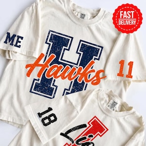 May include: Two off-white short-sleeve t-shirts. The top shirt features a large navy blue distressed letter H with the orange script word Hawks, the letters ME on the sleeve, and the number 11. The bottom shirt has the number 18 and a red letter.