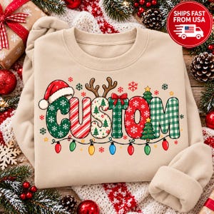 May include: A beige crewneck sweatshirt featuring the word CUSTOM in large, patterned letters. The letters include green with snowflakes, red and white stripes, a tree print, a red bow, red with snowflakes, and green gingham. A string of colorful lights hangs below.