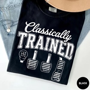 May include: Black t-shirt with the text "Classically TRAINED" in white, along with illustrations of a gear shift and car pedals. The shirt also features the words "Clutch", "Brake", and "Gas".