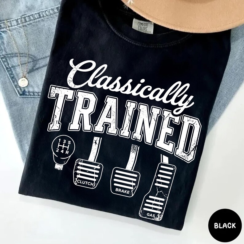 May include: Black t-shirt with the text "Classically TRAINED" in white, along with illustrations of a gear shift and car pedals. The shirt also features the words "Clutch", "Brake", and "Gas".