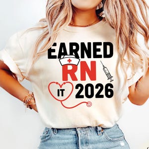 May include: Cream-colored t-shirt with the text "EARNED RN IT 2026". The design includes a nurse's cap, a heart-shaped stethoscope, and a syringe. The text is in black and red.