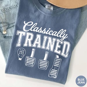 May include: Blue jean colored t-shirt with the words "Classically TRAINED" in white, along with illustrations of a gear shift and car pedals. The shirt is folded on top of a pair of blue jeans.