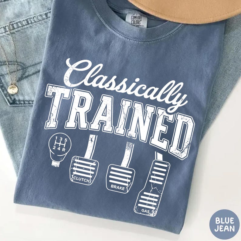 May include: Blue jean colored t-shirt with the words "Classically TRAINED" in white, along with illustrations of a gear shift and car pedals. The shirt is folded on top of a pair of blue jeans.