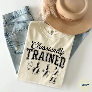 May include: Ivory-colored t-shirt with the text "Classically TRAINED" and illustrations of a gear shift and car pedals. The shirt is paired with light blue jeans and a tan hat.