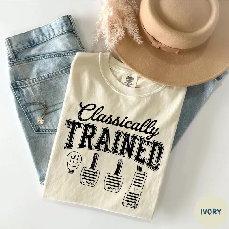 May include: Ivory-colored t-shirt with the text "Classically TRAINED" and illustrations of a gear shift and car pedals. The shirt is paired with light blue jeans and a tan hat.