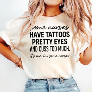 May include: Cream-colored t-shirt with the text "Some nurses have tattoos, pretty eyes, and cuss too much. It's me. I'm some nurses" in black. The shirt is a casual style.