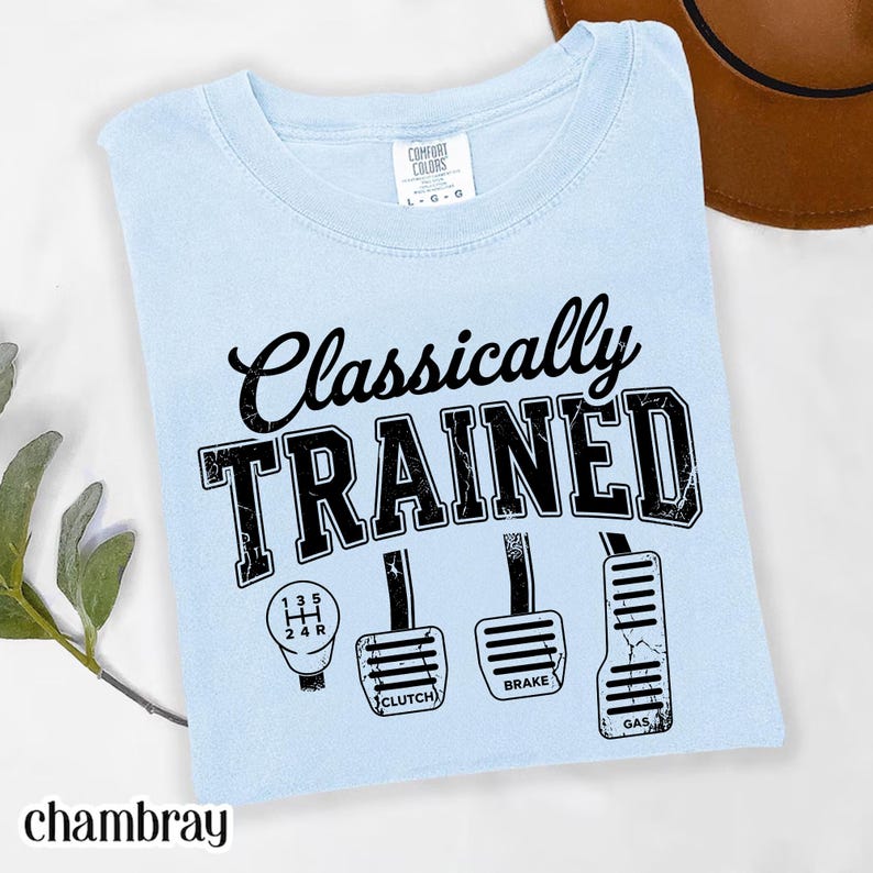 May include: Light blue t-shirt with the words "Classically TRAINED" in black, along with a gear shift and gas, brake, and clutch pedals. The word "chambray" is at the bottom left.