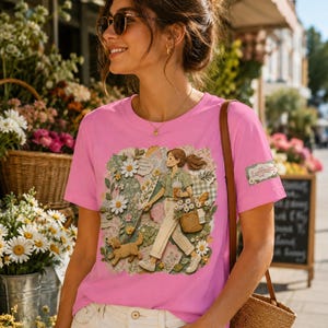 Può includere: Una maglietta rosa con un'illustrazione di una persona e un cane in un giardino, che trasporta un cesto e dei fiori. La maglietta ha una piccola toppa sulla manica con la scritta "Loffeda".