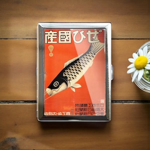 Könnte beinhalten: Eine silberne Zigarettenetui mit einem japanischen Vintage-Design. Das Etui hat einen roten Hintergrund mit einer schwarz-weißen Fischillustration und japanischem Text. Eine Gänseblümchen in einem Glas befindet sich im Hintergrund.