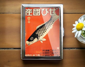 Puede incluir: Un estuche de cigarrillos de metal plateado con un diseño japonés vintage. El estuche presenta un fondo rojo con una ilustración de pez en blanco y negro y texto japonés. Una margarita en un vaso está en el fondo.