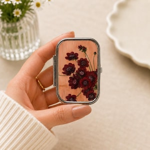 May include: A rectangular metal pill case with a hinged lid featuring a pressed floral design. The lid displays a cluster of deep burgundy flowers with green stems set against a light, natural wood-grain background. The case has a polished silver frame.