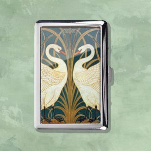 May include: A silver cigarette case with an Art Nouveau design. It shows two white swans, facing each other, set against a teal backdrop with gold and brown plant motifs. The swans have orange beaks and black feet.