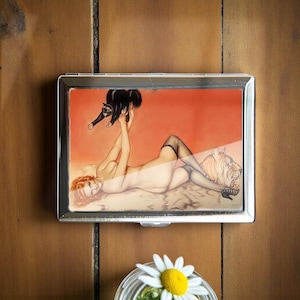 Puede incluir: Un estuche de cigarrillos de metal plateado con una ilustración de estilo pin-up. La obra de arte representa a una mujer pelirroja, un gato negro y un tigre. El fondo es de color naranja cálido. El estuche es rectangular y tiene una tapa con bisagras.