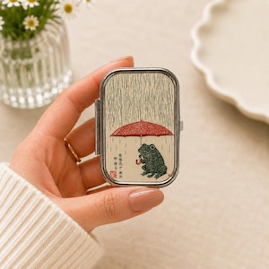 May include: A rectangular metal pill case featuring a printed illustration of a green frog holding a red umbrella under falling rain. The design includes Japanese text and a small red stamp. The case has a silver-toned metal frame and hinge.