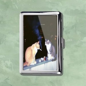 May include: A silver metal cigarette case with a photograph of two cats. One orange tabby cat is smoking a cigarette, and the other cat is gray and white. The cats are sitting on a ledge at night.