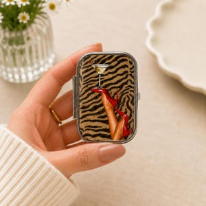 Puede incluir: Un pastillero rectangular de metal con un estampado gráfico de una copa de martini equilibrada sobre un par de piernas con zapatos de tacón rojos sobre un fondo de rayas de cebra marrones y negras. La caja tiene un marco de metal plateado.