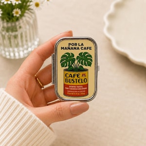 Puede incluir: Una lata metálica rectangular con tapa abatible que presenta un gráfico de una planta monstera creciendo de una lata de café Cafe Bustelo. El texto dice POR LA MAÑANA CAFE, CAFE BUSTELO, SIEMPRE FRESCO, PURO Y AROMATICO COMO NINGUNO, PESO NETO 10 OZ. (283g).