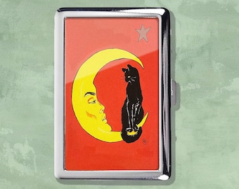May include: A silver metal cigarette case with a red background featuring a yellow crescent moon with a face and a black cat. A star is in the upper right corner. The case has a hinged opening and a silver frame.