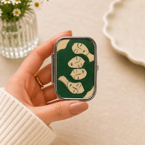 May include: A rectangular metal pill box featuring a painted illustration of four faces with closed eyes and dark green hair. The case has a silver-toned metal frame with a hinged lid and a secure clasp mechanism on the side for opening and closing.