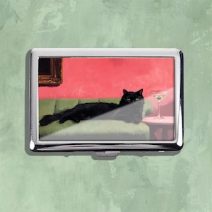 Könnte beinhalten: Eine silberne Zigarettenetui mit einer Illustration einer schwarzen Katze, die sich auf einem grünen Sofa entspannt, einem Martini auf einem Beistelltisch und einer roten Wand im Hintergrund. Es gibt ein gerahmtes Bild.